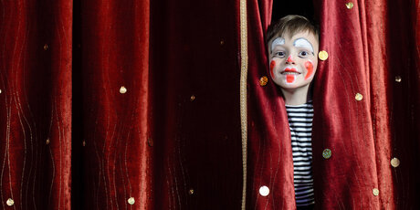 Child made up as a clown, behind a red curtain 