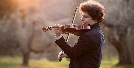 Augustin Hadelich spielt im freien Geige, Bäume im Hintergrund
