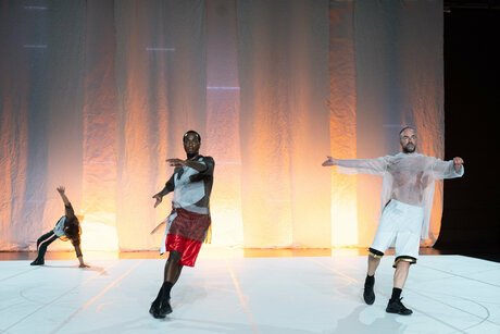Drei Tänzer:innen in lockerer Sportbekleidung auf einer Bühne. Die Person im Hintergrund befindet sich in einer Stützposition.