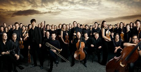 Gruppenbild des Orchesters Mahler Chamber Orchestra vor dramatischem Himmel