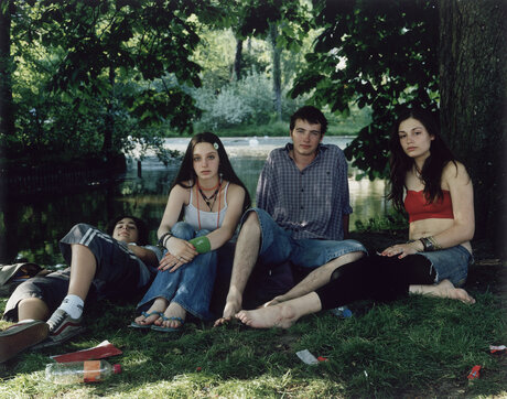 Fotografie: Vier junge Personen sitzen bzw. liegen in Sommerkleidung in einem Park. Ihr direkt ist direkt in die Kamera gerichtet.