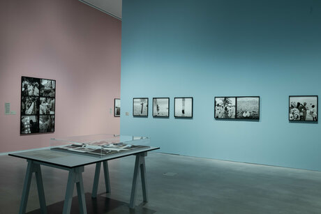 In einer gläsernen Auslage in der Mitte des Ausstellungsraums liegen Fotobücher. Im Hintergrund sind verschiedene Schwarz-Weiß-Fotografien an den pastel-rosa und pastel-blauen Ausstellungswänden zu sehen.