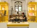 Zwischen zwei gelben Vitrinen mit unterschiedlichen Objekten blickt man auf einen Tisch mit zahlreichen, verschiedenen Goldschmiede-Geräten. An dem Tisch sitzt eine Frau und arbeitet. Zwischen zwei gelben Vitrinen mit unterschiedlichen Objekten blickt man auf einen Tisch mit zahlreichen, verschiedenen Goldschmiede-Geräten. An dem Tisch sitzt eine Frau und arbeitet.