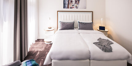 Ein modernes Hotelzimmer mit einem Doppelbett, zwei Nachttischen und einem teilweise geöffneten, deckenhohen Fenster mit Vorhängen. An der Wand über dem Bett ist ein Foto von einem Boot angebracht, und auf dem Bett liegt eine Decke.
