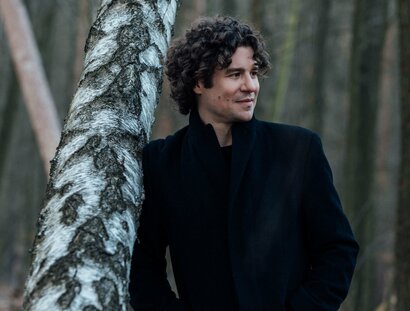Robin Ticciati lehnt im Mantel an einem Baum im Wald