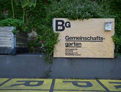 Foto: Ein Holz-Schild trägt die Aufschrift „Gemeinschaftsgarten“ und das Logo der Berlinischen Galerie. Um das Schild herum wachsen Pflanzen. Links daneben ist ein weiterer Pflanzkübel zu sehen.