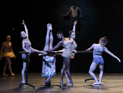 Eine Gruppe von Tänzer:innen trainiert an einer Ballettstange. Eine Person steht auf dem Kopf.