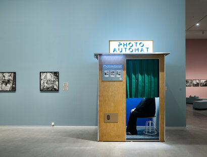 Ausstellungsansicht „Akinbode Akinbiyi“: Blick auf einen Photo-Apparat, in dem eine Person hinter einem dunkelgrünen Vorhang sitzt. Links neben dem Automaten hängen zwei Schwarz-Weiß-Fotografien. Rechts neben dem Automaten ist ein weiterer Ausstellun