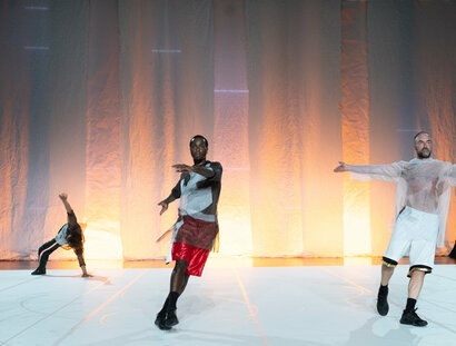 Drei Tänzer:innen in lockerer Sportbekleidung auf einer Bühne. Die Person im Hintergrund befindet sich in einer Stützposition.