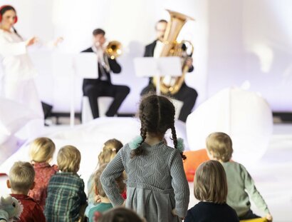 Kinder im Publikum, zwei Musiker spielen Musik, zwei Frauen in weißen Anzügen