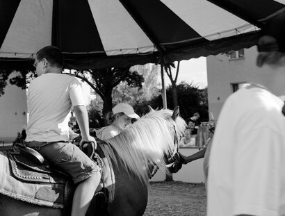 Schwarz-Weiß-Fotografie: Seitenansicht von einem Jungen auf einem Pferd unter einem gestreiften Baldachin. Im Hintergrund sind weitere Menschen zu sehen.