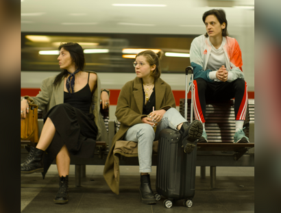 Drei Personen sitzen mit gepackten Koffern am Bahnhof. Im Hintergrund sieht man einen durchfahrenden Schnellzug.