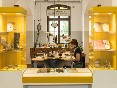 Zwischen zwei gelben Vitrinen mit unterschiedlichen Objekten blickt man auf einen Tisch mit zahlreichen, verschiedenen Goldschmiede-Geräten. An dem Tisch sitzt eine Frau und arbeitet.