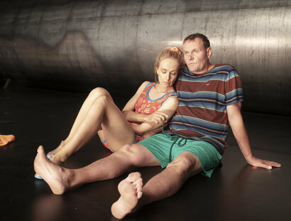 Eine Frau und ein Mann in sommerlicher Kleidung sitzen auf dem Boden. Dahinter ein riesiges Metallrohr.