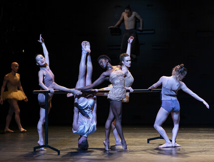 Eine Gruppe von Tänzer:innen trainiert an einer Ballettstange. Eine Person steht auf dem Kopf.