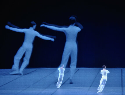 Zwei Tänzer:innen auf einer leeren Bühne. Im Hintergrund eine Projektion der zwei Tänzer:innen.