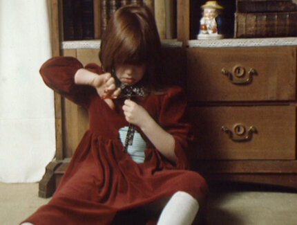 Ein kleines Mädchen sitzt vor einem Schrank. Sie trägt ein rotes Kleid, in das sie sich mit einer Schere Löcher schneidet.