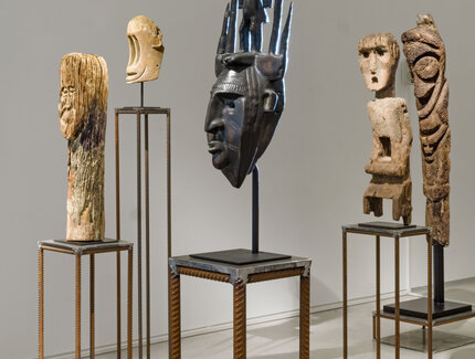 Foto: Blick auf fünf Stehlen aus geriffeltem Stahl, auf denen je eine skulpturartige Darstellung eines Gesichts aus je verschiedenen Materialien angebracht ist.