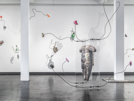 Blick in einen Ausstellungsraum, in dem eine Installation aus dünnen, gebogenen Metallstangen zu sehen ist, an die bunte Objekte angebracht sind.
