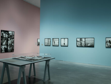 In einer gläsernen Auslage in der Mitte des Ausstellungsraums liegen Fotobücher. Im Hintergrund sind verschiedene Schwarz-Weiß-Fotografien an den pastel-rosa und pastel-blauen Ausstellungswänden zu sehen.