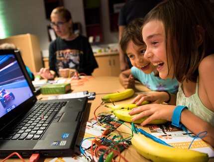 Kinder sitzen an einem Tisch vor einem Laptop. Auf dem Bildschirm ist ein Spiel mit einem Rennauto zu sehen. Die Kinder steuern das Spiel mit Bananen, die auf dem Tisch liegen und über Kabel und ein Gerät mit dem Laptop verbunden sind.