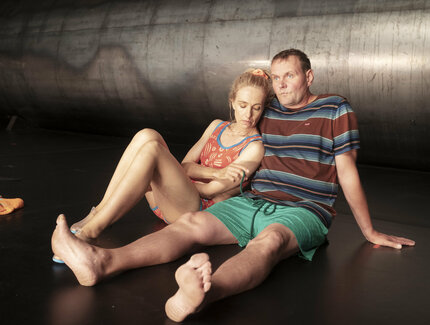 Eine Frau und ein Mann in sommerlicher Kleidung sitzen auf dem Boden. Dahinter ein riesiges Metallrohr.