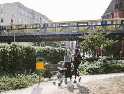 Young man carrying daughter on shoulders while woman pushing baby stroller at footpath against railway bridge