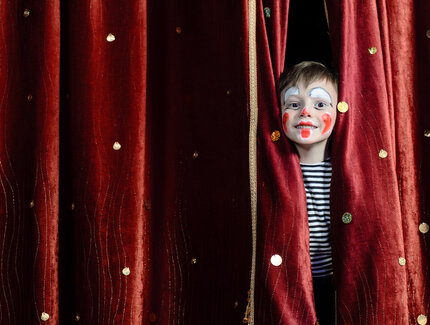 Child made up as a clown, behind a red curtain 